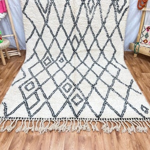May include: A white and black Moroccan rug with a diamond pattern and fringe. The rug is made of wool and has a soft, fluffy texture.