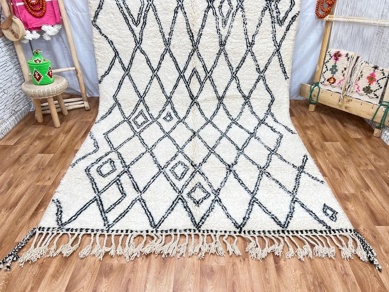 May include: A white and black Moroccan rug with a diamond pattern and fringe. The rug is made of wool and has a soft, fluffy texture.