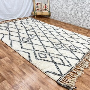 May include: A white and black Moroccan rug with a diamond pattern and fringe. The rug is laid out on a wooden floor.