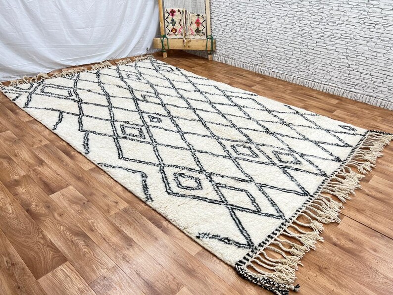 May include: A white and black Moroccan rug with a diamond pattern and fringe. The rug is laid out on a wooden floor.