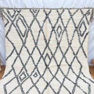 May include: A white and black Moroccan rug with a diamond pattern. The rug has a fringe border and is made of wool.