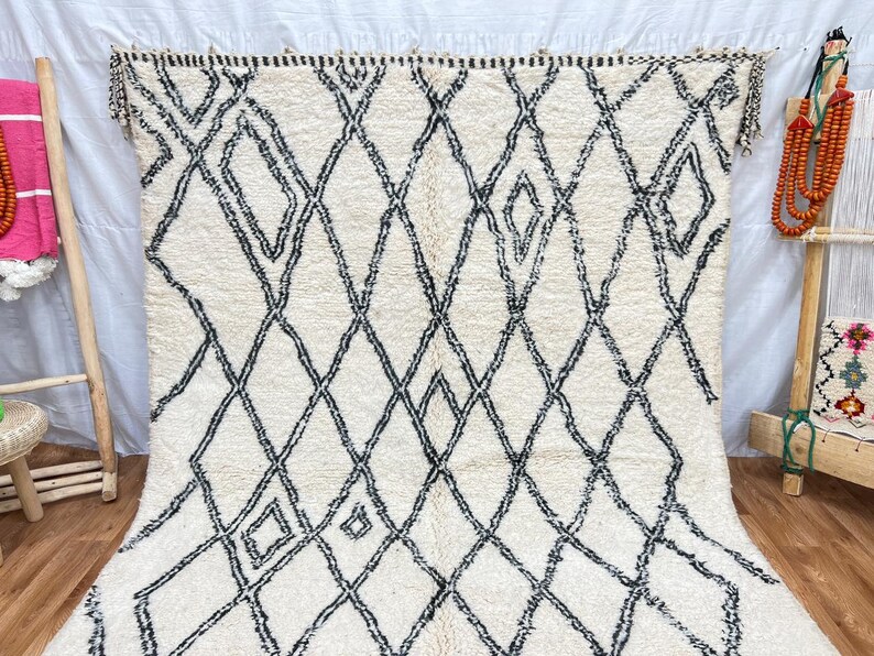 May include: A white and black Moroccan rug with a diamond pattern. The rug has a fringe border and is made of wool.