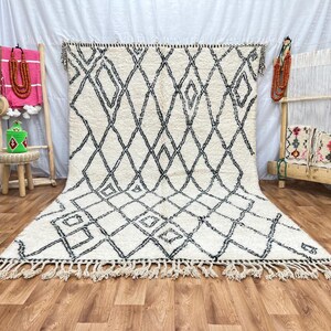 May include: A white and black Moroccan rug with a diamond pattern. The rug has a fringe border and is laid out on a wooden floor.