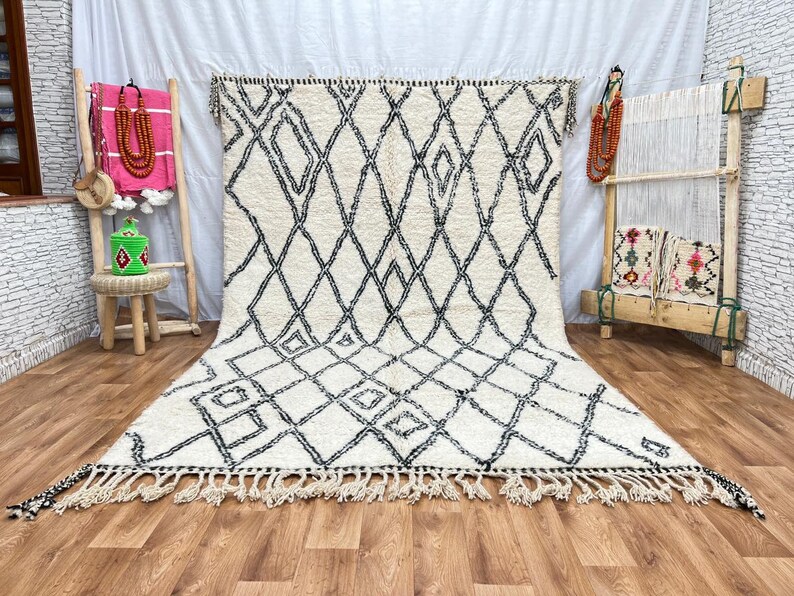 May include: A white and black Moroccan rug with a diamond pattern. The rug has a fringe border and is laid out on a wooden floor.