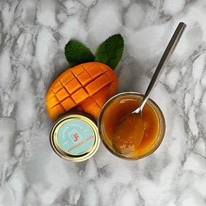 May include: A glass jar of mango jam with a silver spoon inside. The jar is on a white marble surface next to a slice of mango and a small jar with a label that reads "Jammin Fruits Artisan Jams".
