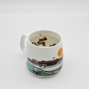 May include: White ceramic mug with a colorful image of the Potala Palace in Lhasa, Tibet. The mug is filled with a light brown herbal tea.