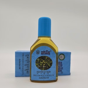 May include: A blue bottle of Drumm Bue Juknuum Arthritis Oil with a blue cap and a label with the text "གྲུམ་བུའི་བྱུག་སྣུམ།" and "DRUUM BUE JUKNUUM Arthritis Oil".