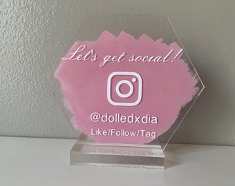 May include: A pink hexagonal acrylic sign with the text "Let's get social!" and an Instagram logo. The sign also includes the text "@dolledxdia" and "Like/Follow/Tag".