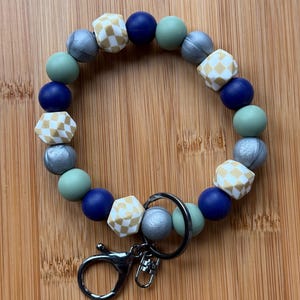 May include: A silicone beaded keychain with a silver, blue, and green color scheme. The beads have a geometric pattern and are strung on a silver ring with a silver clip.