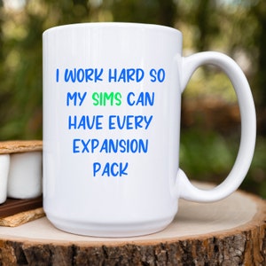 May include: White ceramic coffee mug with blue text that reads "I work hard so my Sims can have every expansion pack".