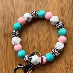 May include: A pink, turquoise, and white beaded keychain with a silver key ring. The beads are made of silicone and have a variety of patterns, including marble, cow print, and solid colors.