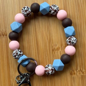 May include: A silicone bracelet with a key chain attachment. The bracelet is made of brown, pink, blue, and white beads. The beads have a variety of patterns, including cow print and solid colors.