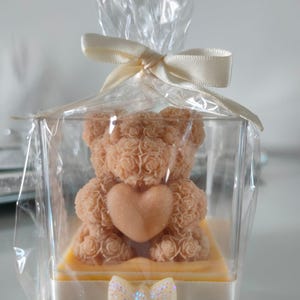 Op de afbeelding: Een lichtbruine teddybeer-vormige zeep met een hart, verpakt in een transparante doos. De beer staat op een gele basis en is verpakt in cellofaan, vastgebonden met een crèmekleurig lint en een glitterstrik. Een charmant cadeau-idee.