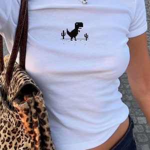 May include: White crew neck t-shirt with a black pixelated dinosaur and cacti design. The shirt is worn with dark blue denim jeans and a leopard print handbag. A silver necklace is also visible.