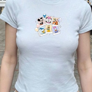May include: Pale blue t-shirt with a collection of Disney character images in a polaroid style. The images feature Mickey Mouse, Minnie Mouse, Goofy, Donald Duck, Pluto, and Daisy Duck. The top has short sleeves and a round neck.