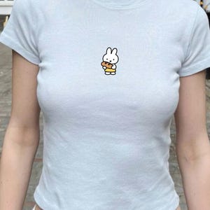 May include: Pale blue t-shirt with a cartoon bunny graphic. The bunny is white with black details, holding a brown loaf. The t-shirt has a round neck and short sleeves.
