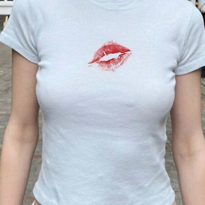 May include: Pale blue t-shirt featuring a red lipstick kiss print. The short-sleeved top has a scoop neck and is made of a soft fabric. The shirt is worn with white trousers.