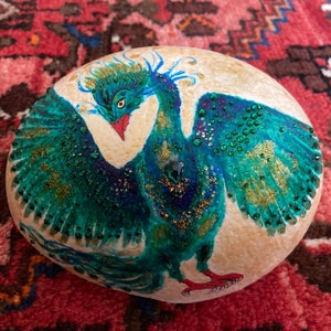 Könnte beinhalten: Ein handbemalter Stein mit einem Pfauen-Design. Der Pfau ist in Blautönen, Grün- und Goldtönen mit Glitzerakzenten bemalt. Der Stein ist hellbraun.