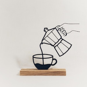 Könnte beinhalten: Schwarz-Weiß-Illustration eines Kaffeezubereiters, der Kaffee in eine Tasse gießt. Die Tasse und der Kaffeezubereiter sind schwarz, und das Bild steht auf einem Holzsockel. Der Hintergrund ist weiß.