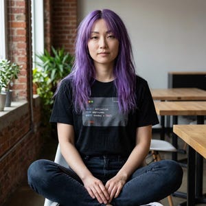 Puede incluir: Una camiseta negra con el texto "SELECT motivation FROM employees WHERE monday = TRUE;" es usada por una persona con cabello morado. La persona está sentada en una silla con las piernas cruzadas. El fondo incluye una pared de ladrillos y una ventana.
