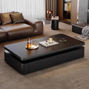 Pode incluir: Uma mesa de centro retangular preta moderna com uma seção de armazenamento inferior. A mesa tem cantos arredondados e uma superfície lisa. Uma bandeja com copos e uma garrafa está em cima, junto com uma vela e um livro. A mesa está em uma sala de estar.
