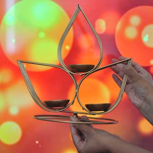 May include: A gold metal candle holder shaped like a lotus flower with three candle cups. The candle holder is being held by two hands against a background of blurred colorful lights.