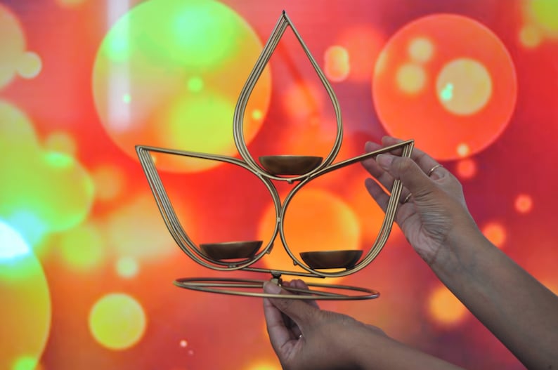 May include: A gold metal candle holder shaped like a lotus flower with three candle cups. The candle holder is being held by two hands against a background of blurred colorful lights.