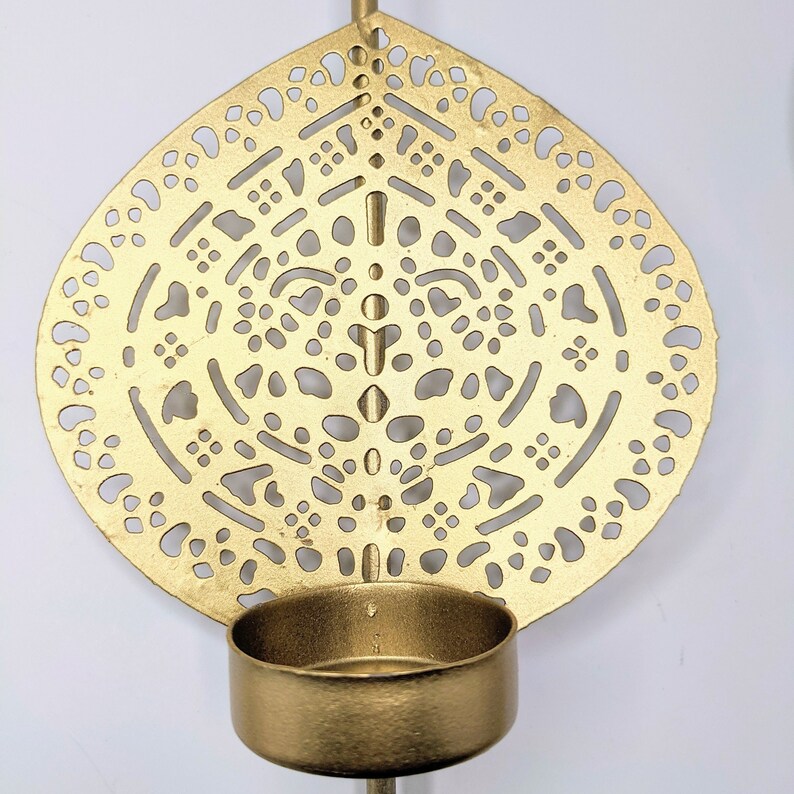 May include: Gold metal candle holder with a decorative, circular, cutout design. The holder has a round, open top for a candle.