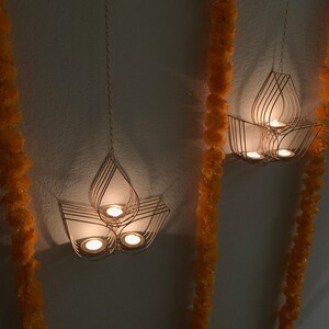 May include: Three gold metal candle holders hanging from the ceiling with lit candles inside. The candle holders are shaped like lotus flowers. The candle holders are hanging in front of a white wall with orange flower garlands on either side.