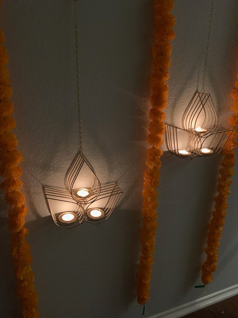 May include: Three gold metal candle holders hanging from the ceiling with lit candles inside. The candle holders are shaped like lotus flowers. The candle holders are hanging in front of a white wall with orange flower garlands on either side.