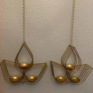 May include: Two gold metal candle holders with a teardrop design. Each holder has three candle cups and hangs from a chain.