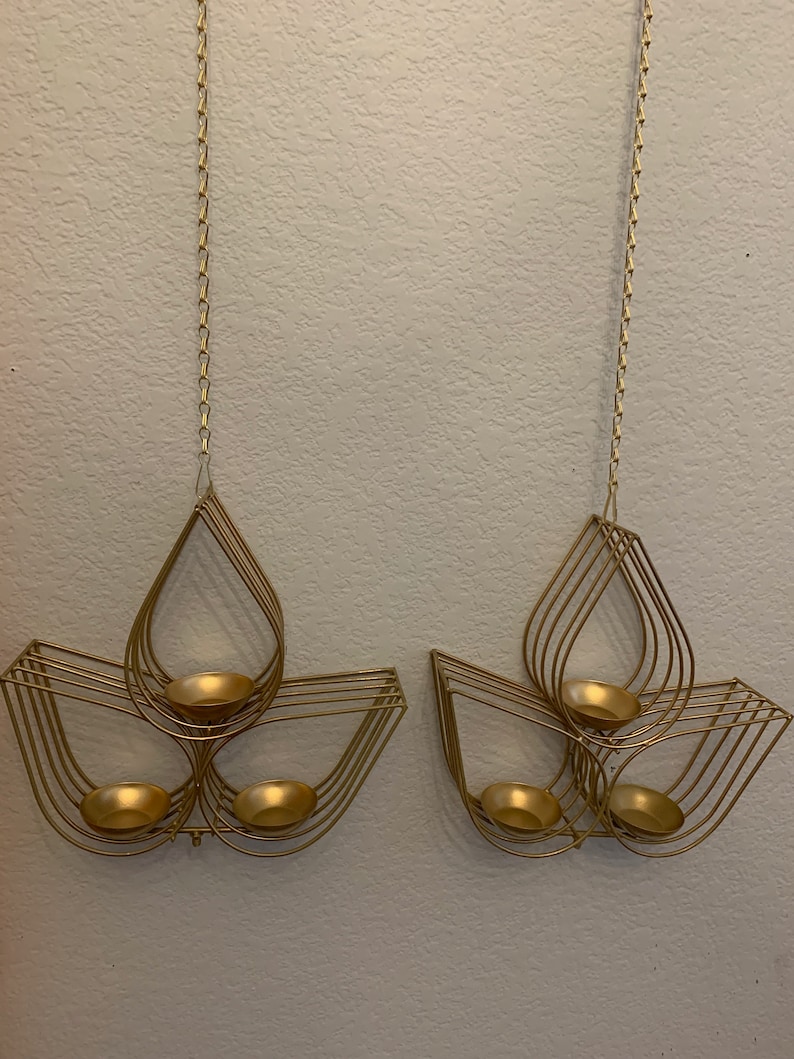 May include: Two gold metal candle holders with a teardrop design. Each holder has three candle cups and hangs from a chain.