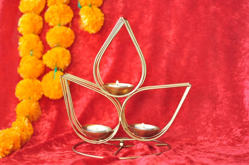 May include: A gold metal candle holder shaped like a lotus flower with three candle cups. The candle holder is on a red velvet surface.