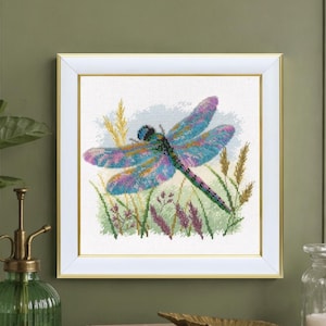 May include: A framed cross-stitch artwork featuring a colorful dragonfly with blue, purple, and pink wings. The dragonfly is set against a backdrop of green grass and wheat-colored stalks. The artwork is in a white frame.