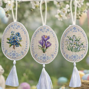 Peut inclure: Trois décorations brodées en forme d'œufs de Pâques avec des motifs floraux. Chaque ornement en forme d'œuf présente une fleur différente : myosotis bleus, crocus violets et perce-neige blancs. Ils sont suspendus à des cordons blancs et ont des glands blancs.