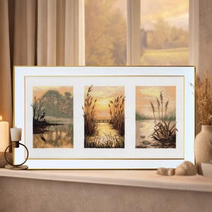 May include: A framed triptych featuring three panels of a serene landscape scene. Each panel depicts a waterside view with reeds, trees, and a sunset. The artwork is framed in white with a gold border, creating a calming aesthetic.