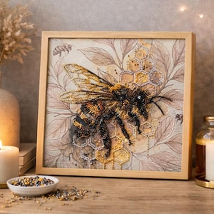 May include: Framed artwork featuring a detailed bee design. The bee is rendered with intricate beadwork in shades of black, gold, and brown, set against a backdrop of a honeycomb and leaves. The artwork is displayed on a wooden surface.