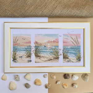 May include: A framed cross-stitch artwork depicting a beach scene with a sunset, seagrass, and a sailboat. The artwork is in a white frame with a gold inner border. Seashells are arranged around the frame.