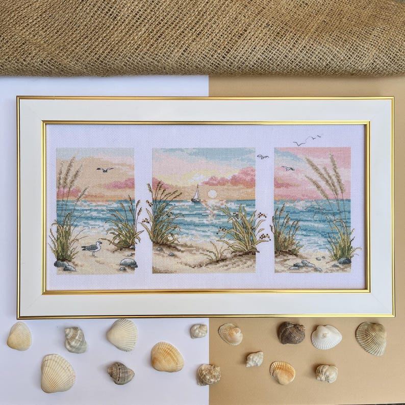 May include: A framed cross-stitch artwork depicting a beach scene with a sunset, seagrass, and a sailboat. The artwork is in a white frame with a gold inner border. Seashells are arranged around the frame.