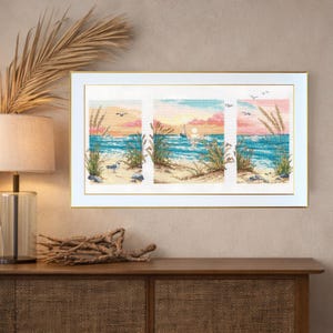 May include: A framed cross-stitch artwork depicting a beach scene with a sunset. The artwork is divided into three panels, showcasing blue water, sand, seagrass, and a sailboat. The frame is white with a gold inner edge. The artwork is displayed on a wooden cabinet.