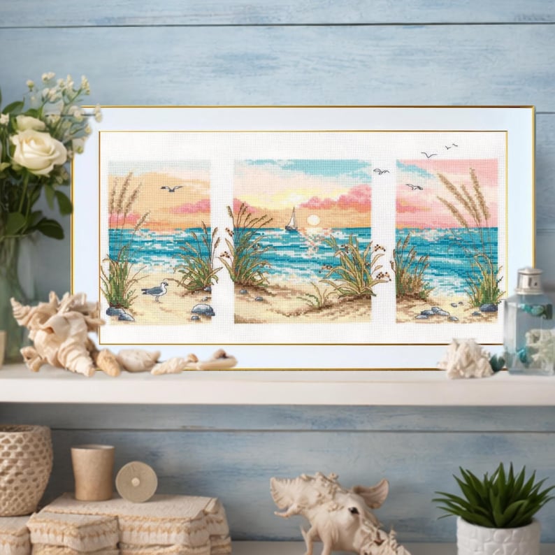May include: Framed cross-stitch artwork depicting a beach scene with a sunset. The artwork features a sailboat, seagulls, and beach grass. The color palette includes shades of blue, pink, and yellow. The frame is white with a gold border.