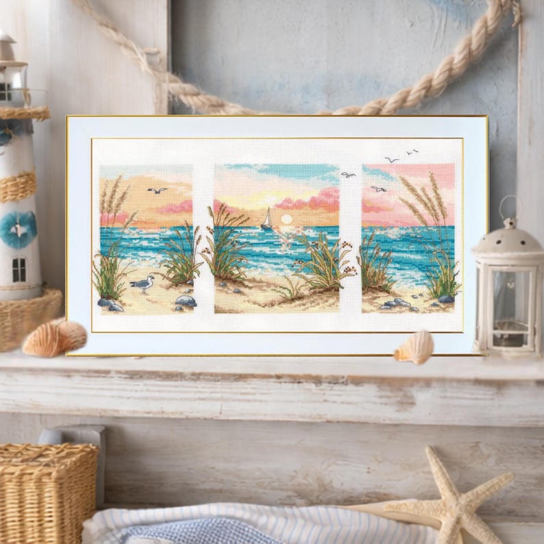May include: A framed cross-stitch artwork depicting a beach scene with a sailboat, seagull, and tall grass. The artwork features a sunset with pink and orange hues over the ocean. The frame is white with a gold trim.
