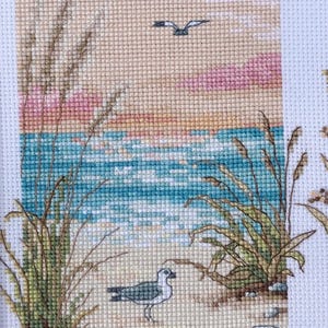 May include: Cross-stitch artwork depicting a serene beach scene. The artwork features a seagull on a sandy shore, with ocean waves, tall grass, and a sunset in the background. The color palette includes shades of blue, beige, pink, and green.