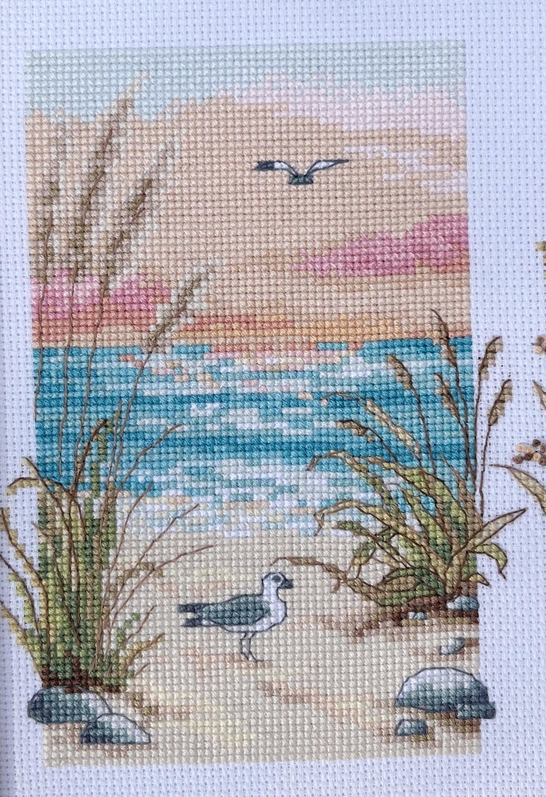 May include: Cross-stitch artwork depicting a serene beach scene. The artwork features a seagull on a sandy shore, with ocean waves, tall grass, and a sunset in the background. The color palette includes shades of blue, beige, pink, and green.