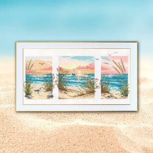 May include: A framed cross-stitch artwork depicting a beach scene. The artwork is divided into three panels, each showcasing a section of the beach with the ocean, seagrass, and a sunset in shades of pink, orange, and blue. A sailboat is visible.