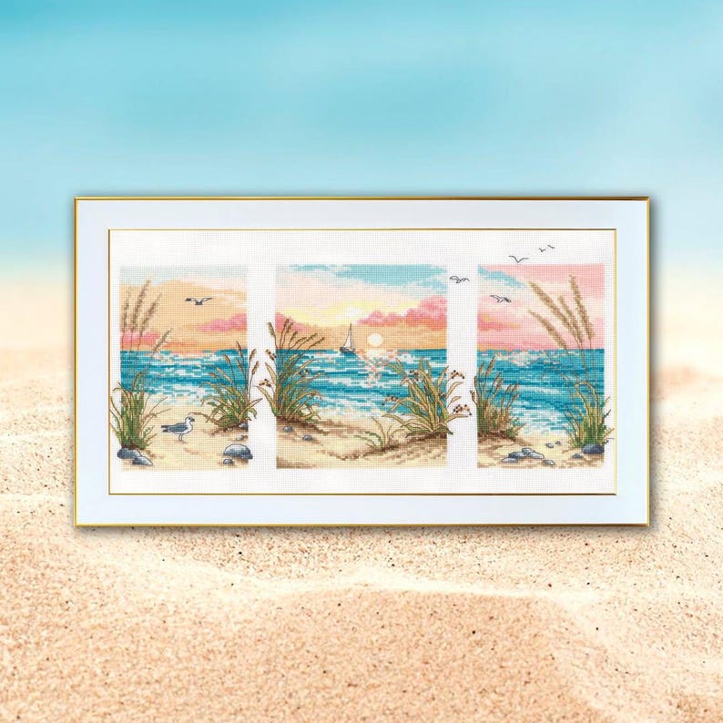 May include: A framed cross-stitch artwork depicting a beach scene. The artwork is divided into three panels, each showcasing a section of the beach with the ocean, seagrass, and a sunset in shades of pink, orange, and blue. A sailboat is visible.