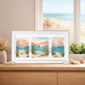 May include: Framed cross-stitch artwork depicting a beach scene with a sunset. The artwork is divided into three panels, showcasing the ocean, sand, and seagrass. The frame is gold, and the artwork is displayed on a wooden surface.