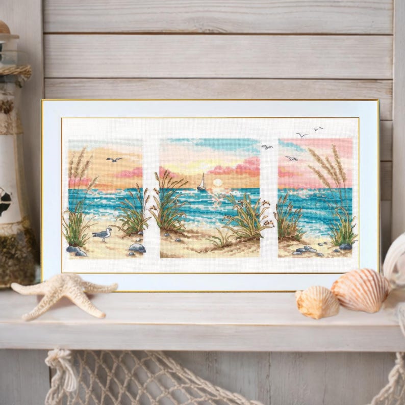 May include: A framed cross-stitch artwork depicting a beach scene with a sailboat, seagull, and tall grass. The artwork features a sunset with pink and orange hues over the blue ocean. The frame is white with a gold inner border.