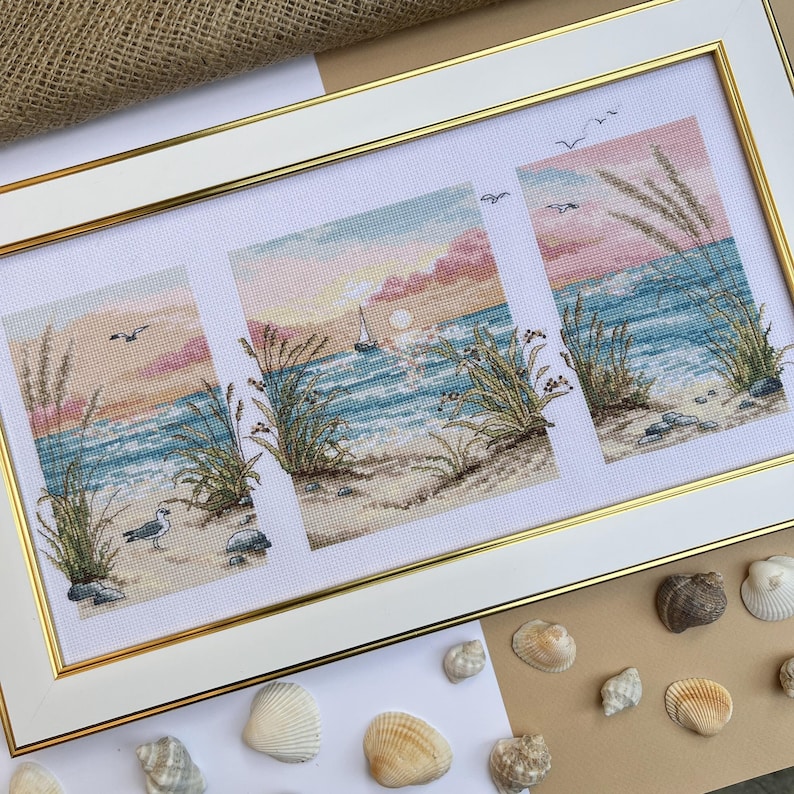 May include: A framed cross-stitch artwork depicting a beach scene with a sunset. The artwork is divided into three panels, each showing waves, sand, and plants. A sailboat is visible in the center panel. The frame is white with a gold inner border.