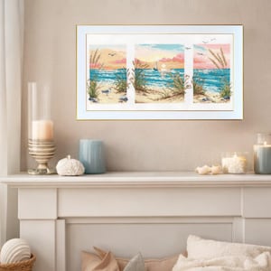 May include: A framed triptych cross-stitch artwork depicting a beach scene. The artwork features a sunset with a sailboat on the ocean, sand, and tall grass. The frame is white with a gold inner edge.